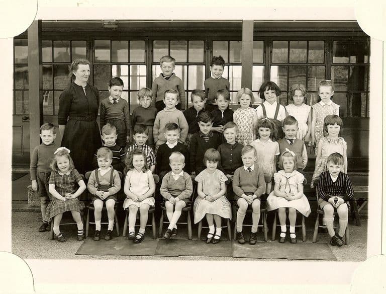 Class of about thirty-five infants in four rows, a woman teacher in a dark dress standing at the left, in front of glazed classroom doors.