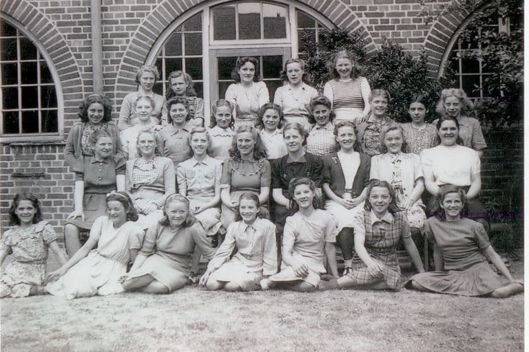 About thirty teenage girls in summer dresses and blouses in four rows, the front row sitting on the grass, in front of a brick school building with large arched windows.