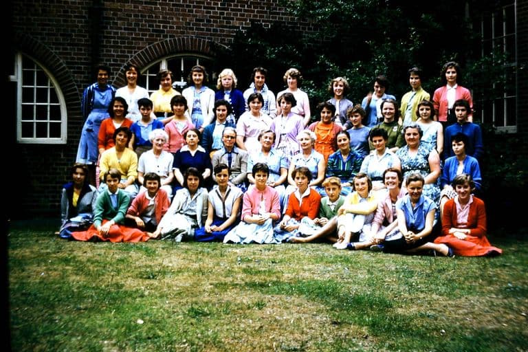 Colour photo of about fifty girls and women teachers in five rows on a lawn in front of a brick school building with an arched window; the girls wear bright cardigans and skirts.