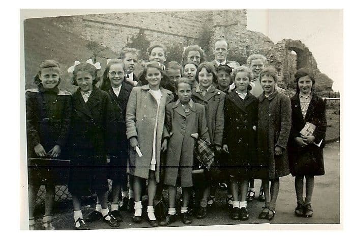 About twenty girls in winter coats and white socks, some with hair ribbons, stand in a group with a male teacher in front of the ruined stone walls of Dudley Castle.