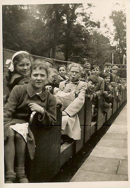 Girls and boys crowded into the open carriages of a miniature railway beside a paved platform, a girl with a large white hair bow and a boy in glasses at the front, trees behind.