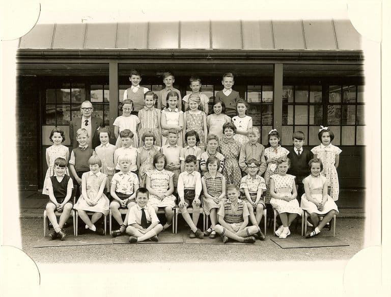 Large class of about forty-five junior-school children in five rows on the playground, a male teacher in glasses standing at the left, in front of a veranda with glazed doors under a pitched canopy.