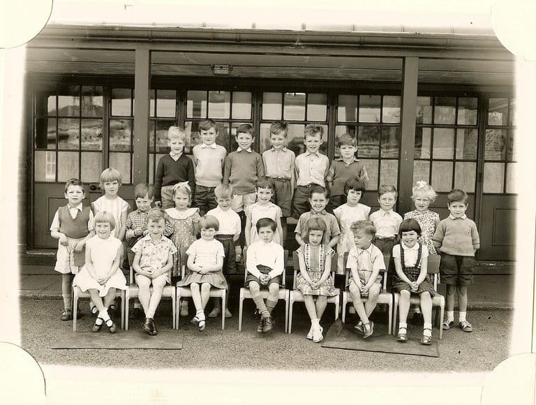 Class of about thirty infants in four rows, the front row on small chairs on mats, in front of a veranda with glazed doors; no teacher appears.