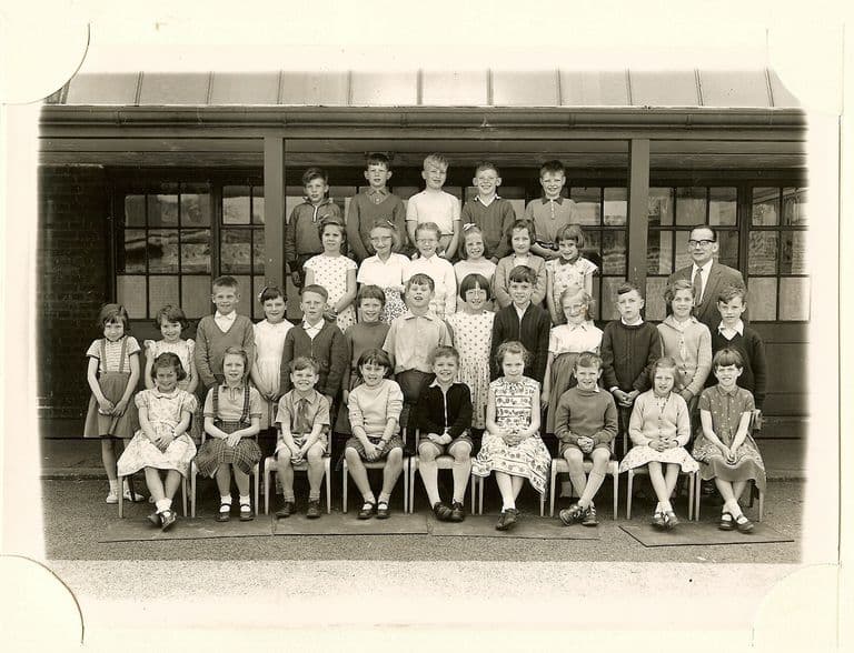 Class of about forty-five children in five rows, a male teacher in glasses standing at the right, in front of a veranda with glazed doors.