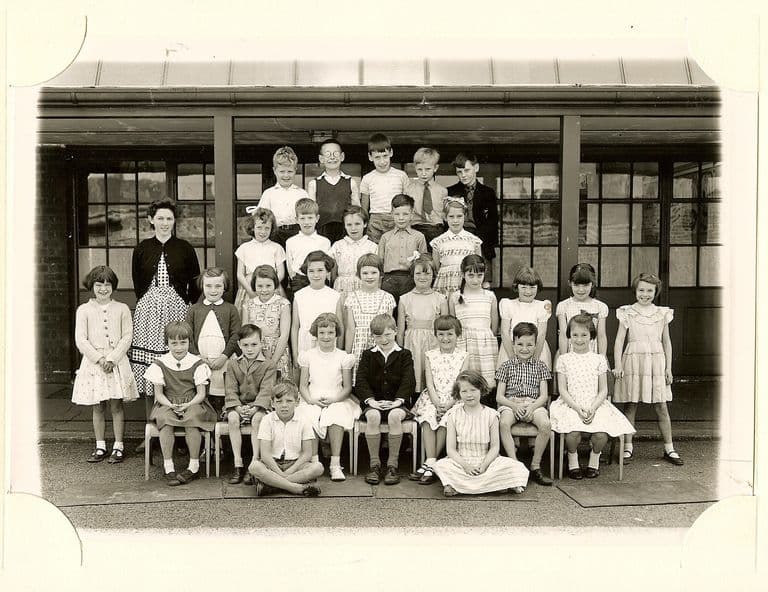 Class of about forty infants in five rows, a woman teacher in a dark cardigan standing at the left, with two children sitting on the ground in front.