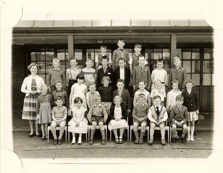 Class of about thirty-five children in five rows, a woman teacher in a light cardigan and patterned skirt standing at the left, in front of glazed classroom doors.