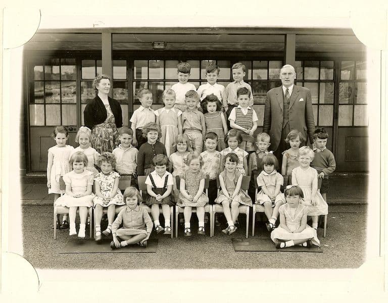 Class of about thirty-five infants in five rows, a woman teacher in a dark cardigan and an older man in a suit standing at the back, two girls sitting on the ground in front.