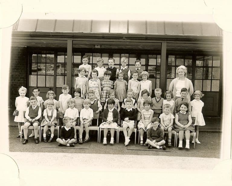 Class of about forty-five infants in five rows, an older woman teacher in a cardigan standing at the right, two boys sitting cross-legged in front.