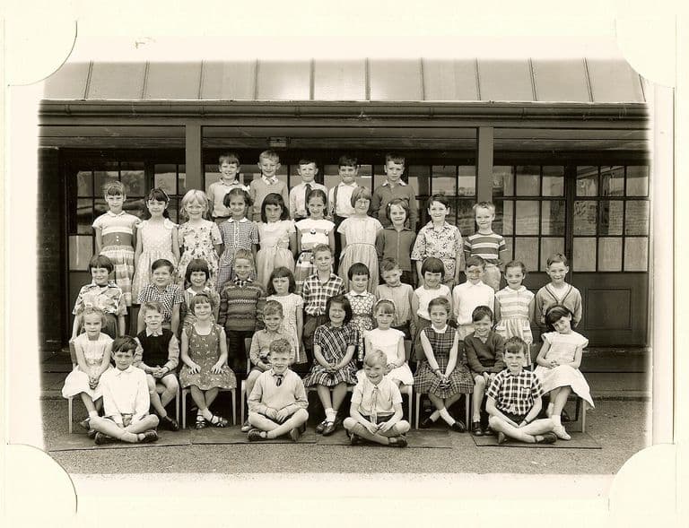 Class of about forty-five children in five rows, four boys sitting cross-legged in front, in front of glazed classroom doors; no teacher appears.