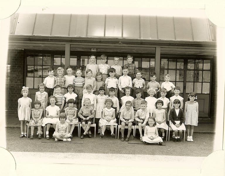 Class of about fifty young children in six rows under a veranda canopy, two girls sitting on the ground in front; no teacher appears.