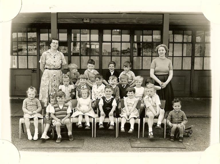 Small group of about twenty very young children with two women teachers, one boy sitting on a toy cart at the right, in front of glazed classroom doors.