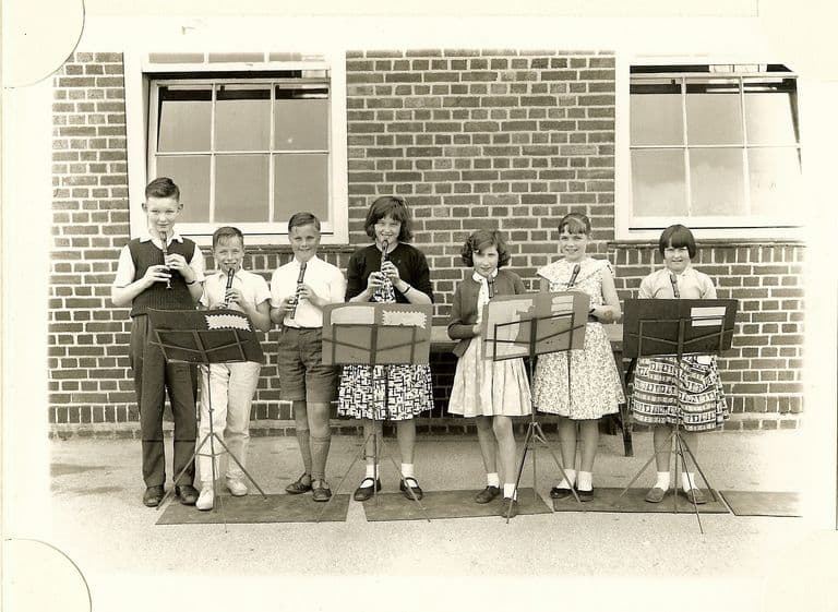 Seven children playing descant recorders at music stands in a row in front of a brick school wall with sash windows.