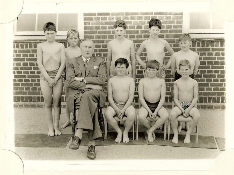 A school team photograph of eight boys in swimming trunks with a headmaster in a suit seated among them, in front of a brick wall.