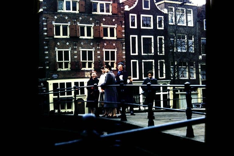 Colour slide of a small group of women and a boy standing on a canal bridge in front of tall gabled Amsterdam houses with shuttered windows.