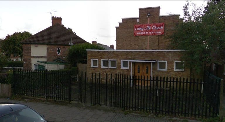 A street view of a brick church hall behind black railings: a single-storey front with a double door and a taller gabled hall behind, hung with a red banner reading “Living Life Church – Sundays at 10.30–12.00”, beside a pair of semi-detached houses. “© 2009 Google” appears faintly on the image.