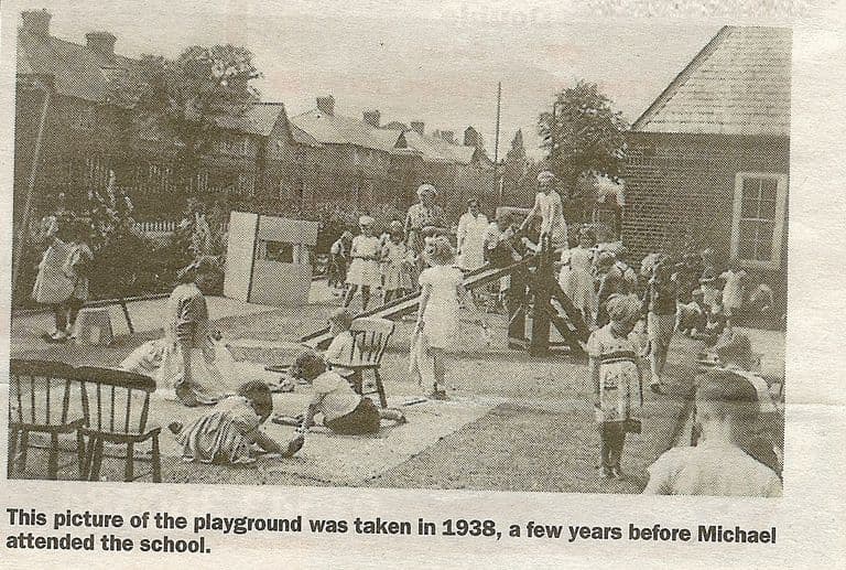 A newspaper photograph of young children playing in a school playground with a seesaw, a small wooden playhouse and chairs, watched by teachers, with houses behind. The printed caption reads “This picture of the playground was taken in 1938, a few years before Michael attended the school.”