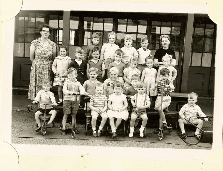 About thirty very young children in four rows with two women teachers, several sitting on tricycles, scooters and a toy car at the front, in front of glazed classroom doors.