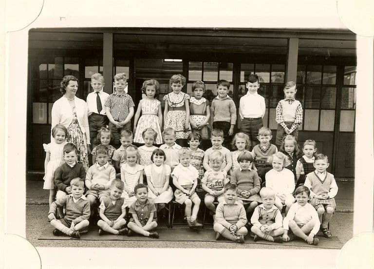 Class of about forty infants in four rows, a woman teacher in a cardigan standing at the left, the front row sitting cross-legged on a mat.