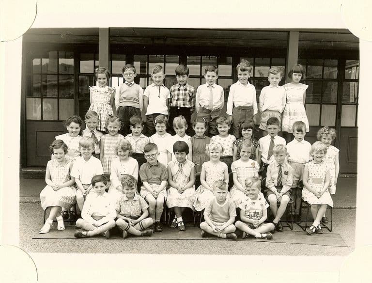 Class of about forty infants in four rows, the front row sitting cross-legged on a mat, in front of a veranda with glazed doors; no teacher appears.
