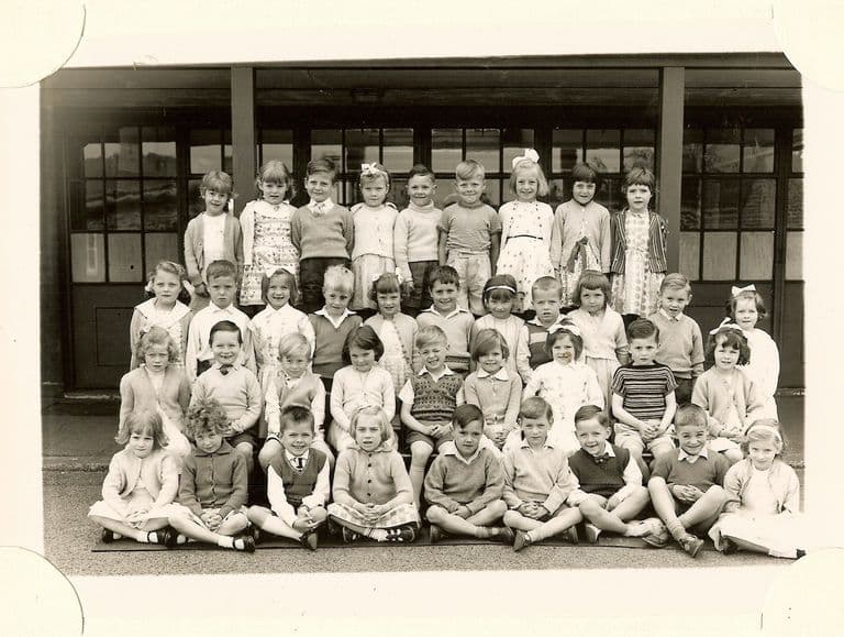 Class of about forty-five infants in four rows, the front row sitting cross-legged, in front of glazed classroom doors; no teacher appears.