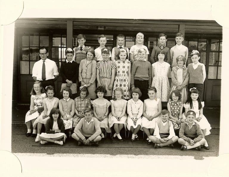 Class of about forty junior-school children in five rows, a young male teacher in a white shirt and tie standing at the left.