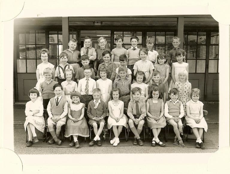Class of about forty children in four rows, a woman teacher in glasses standing at the left, in front of a veranda with glazed doors.