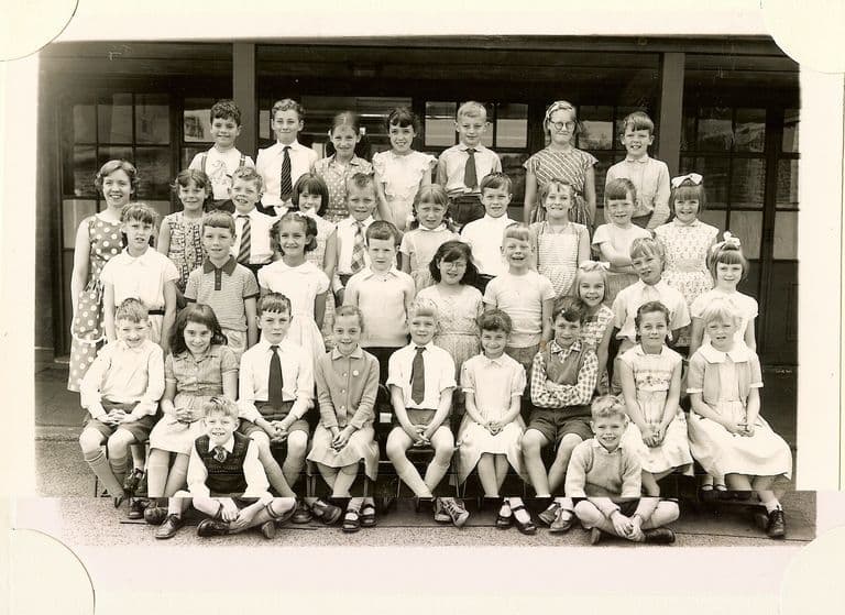 Class of about forty-five junior-school children in five rows, a young woman teacher standing at the left, two boys sitting cross-legged in front.