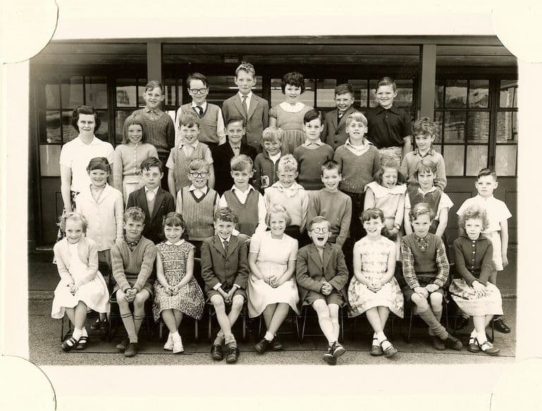 Class of about forty junior-school children in four rows, a woman teacher in a white blouse standing at the left, in front of glazed classroom doors.