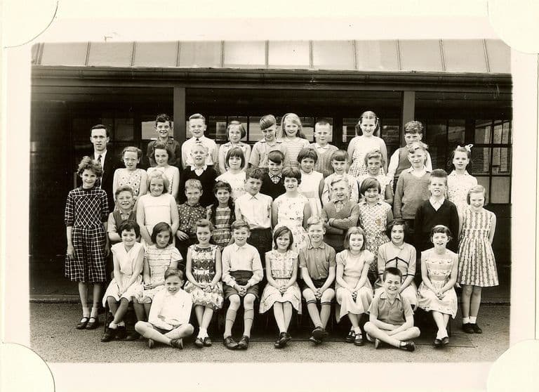 Large class of about fifty junior-school children in six rows under a veranda canopy, a young male teacher standing at the back left.