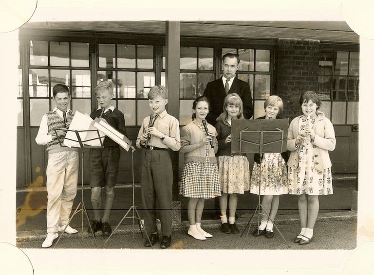 Seven children playing recorders at music stands, with a male teacher in a dark suit standing behind them, in front of glazed classroom doors.