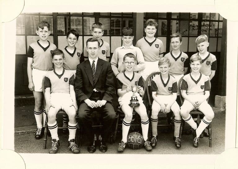 School football team of eleven boys in dark shirts with light collars and hooped socks, one holding a trophy cup, with a young male teacher in a suit; the ball is marked “1961-62”.