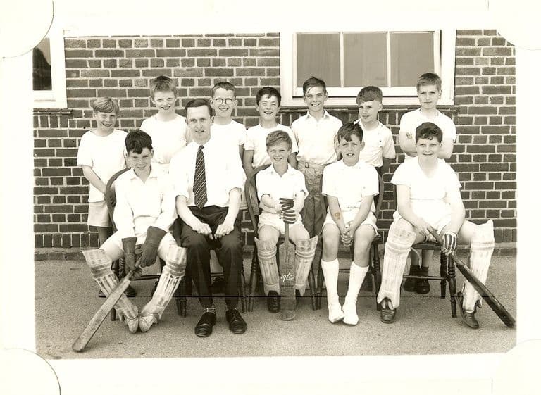 School cricket team of about twelve boys in whites, three with pads and bats, with a young male teacher in a shirt and tie seated in the centre, in front of a brick wall.