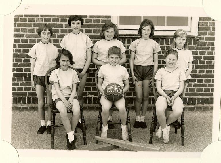 Girls’ sports team of eight girls in light shirts and dark shorts in front of a brick wall, one holding a ball marked “1961-62”, with a bat on the ground in front.