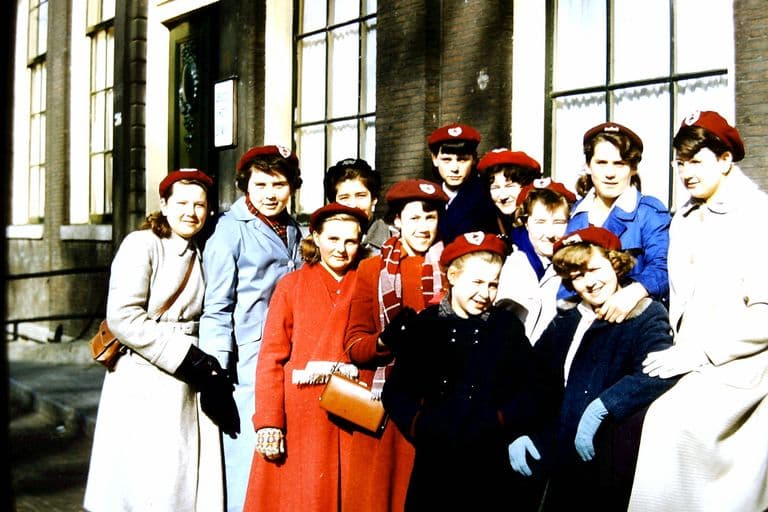Colour slide of about a dozen teenage girls in matching red berets with badges and winter coats, smiling in a huddle outside the tall windows of a brick hotel.