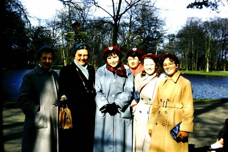 Colour slide of two older women teachers in winter coats and four girls in red berets and long coats standing beside a lake in the parkland at Keukenhof, a signpost behind them.