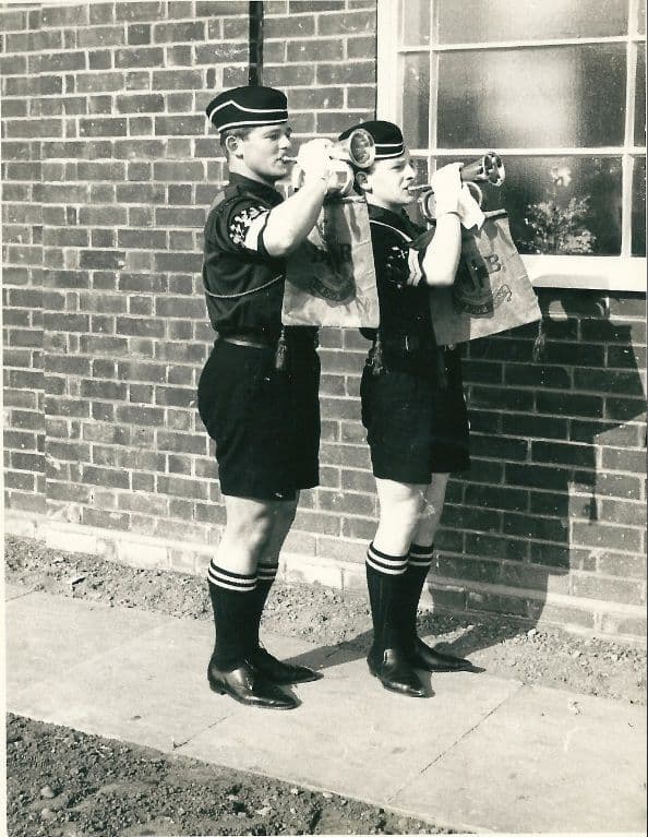 Two Boys’ Brigade buglers in pillbox caps, dark shirts, shorts and striped socks sound bugles with BB banners against the brick wall of a new building.