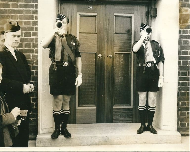 Two Boys’ Brigade buglers in uniform sound bugles either side of the panelled double doors of a new church, while an officer in a glengarry cap and onlookers watch at the left.