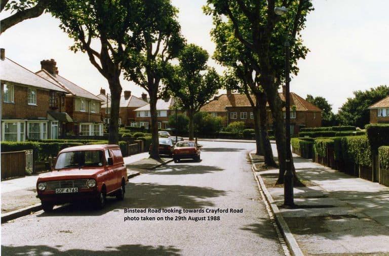 Colour photo of a tree-lined Binstead Road curving between brick semi-detached houses with hedges, a red Mini van parked on the left; a caption is printed on the image.