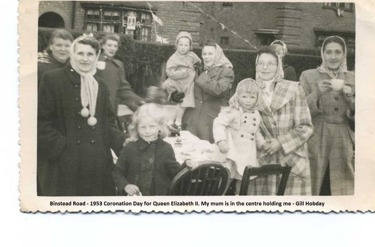 A snapshot of women in headscarves and coats and young children around a table set out in the street, with bunting and a portrait of the Queen with “E R” in a house window behind. A printed caption below reads “Binstead Road – 1953 Coronation Day for Queen Elizabeth II. My mum is in the centre holding me – Gill Hobday”.