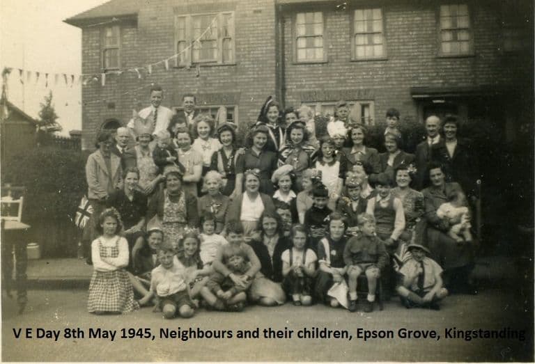 A large group of neighbours – women, men and children, some in paper hats and waving flags – gathered in front of brick houses hung with bunting; a caption is printed on the image.