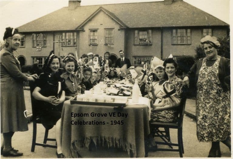 Women and girls in party hats, some holding Union flags, sit at a table in the road with jugs and plates, two older women in aprons standing at either end, in front of rendered semi-detached houses with flags in the windows.