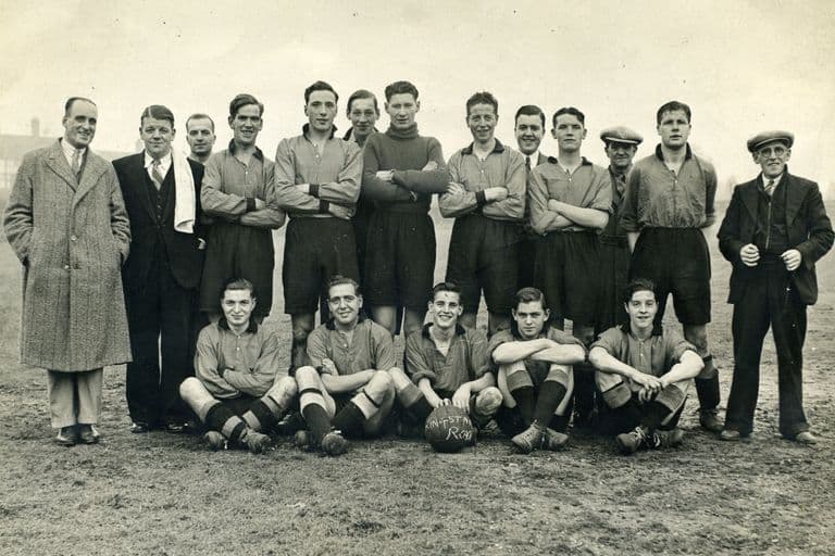 A football team in dark shirts and shorts, the back row standing with arms folded and five players crouching in front around a ball lettered “Kingstanding Royals”, flanked by men in overcoats and caps on a muddy field.
