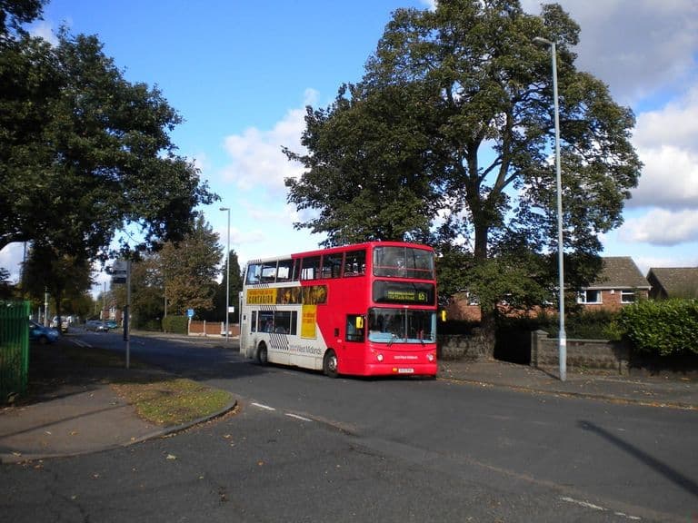 Red and white double-decker bus turning through Court Lane under big trees at the Perry Common bus terminus