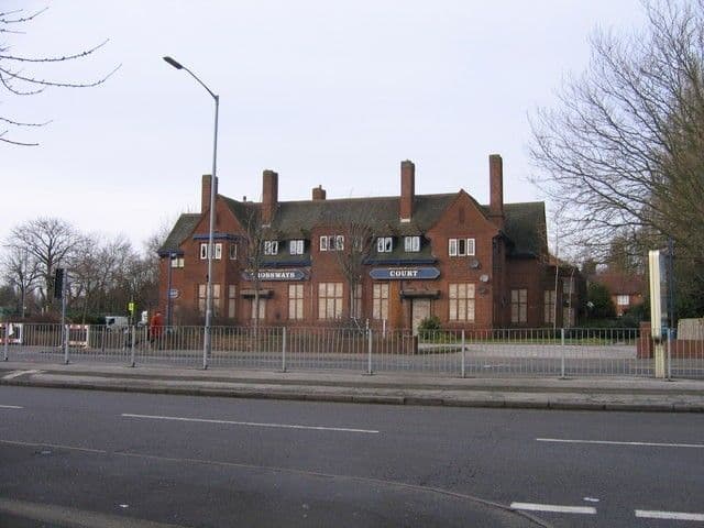 Large brick former pub with tall chimneys and gables, signed 'Crossways Court', standing empty behind railings at a crossroads