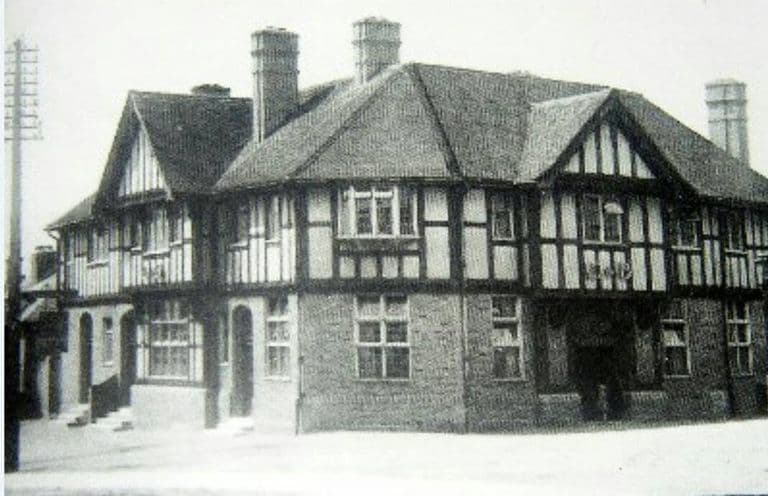 A large mock-Tudor corner building with black-and-white half-timbering, jettied upper floor, tall brick chimneys and a telegraph pole
