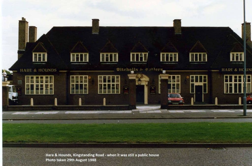 1988: The long front of the Hare & Hounds pub with seven dormer windows, the names Hare & Hounds and Mitchells & Butlers in gold, and a grass verge in front