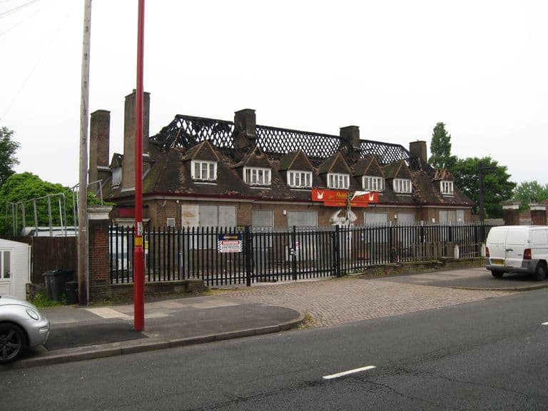 The 1930s Hare and Hounds pub on Kingstanding Road with dormer windows and chimneys, its roof burnt through to bare rafters, behind a fence