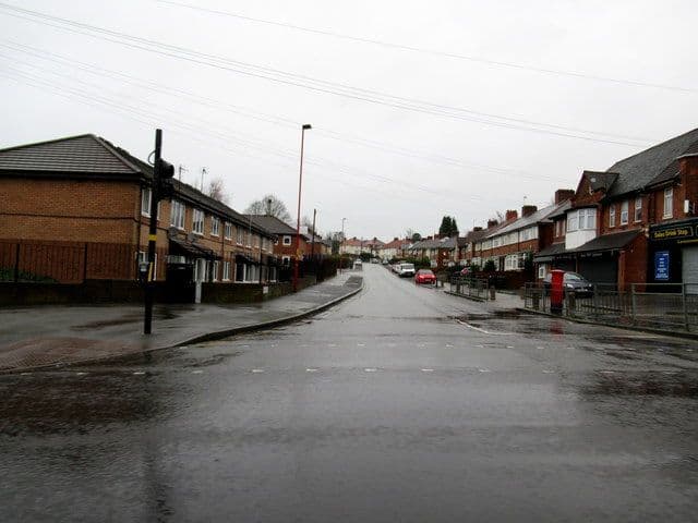 Hartley Road on a wet day, a straight road between terraces of brick houses with a shop and post box on the right.