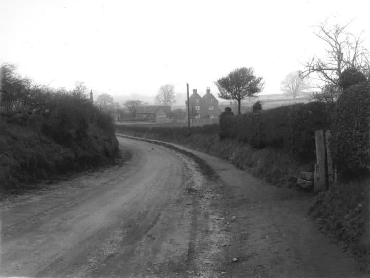 A curving unmade country road between hedges, with a house and trees in the distance.