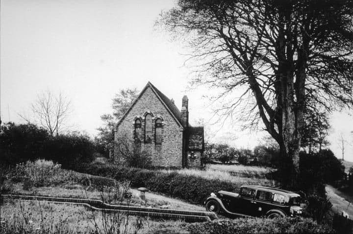1938: A small gabled brick church among trees, with a 1930s car parked on the lane.